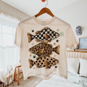 May include: Cream-colored t-shirt featuring three fish with unique patterns: a black and white checkerboard, leopard print, and brown polka dots. The shirt is adorned with stars and shamrocks, creating a playful and eye-catching design. The shirt is hanging on a wooden hanger.