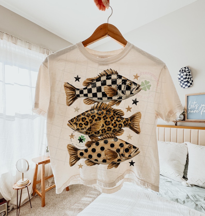 May include: Cream-colored t-shirt featuring three fish with unique patterns: a black and white checkerboard, leopard print, and brown polka dots. The shirt is adorned with stars and shamrocks, creating a playful and eye-catching design. The shirt is hanging on a wooden hanger.