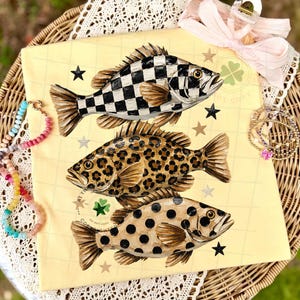 May include: A yellow graphic tee featuring three stylized fish with unique patterns: a black and white checkerboard, leopard print, and brown polka dots. The design includes black stars and a clover emblem.