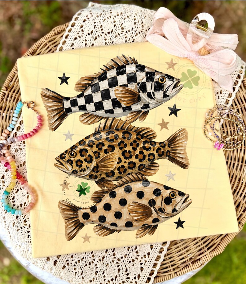 May include: A yellow graphic tee featuring three stylized fish with unique patterns: a black and white checkerboard, leopard print, and brown polka dots. The design includes black stars and a clover emblem.
