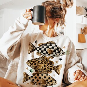 May include: Cream-colored sweatshirt featuring three fish designs: a black and white checkered fish, a leopard print fish, and a fish with brown polka dots. The sweatshirt also has star and clover graphics.