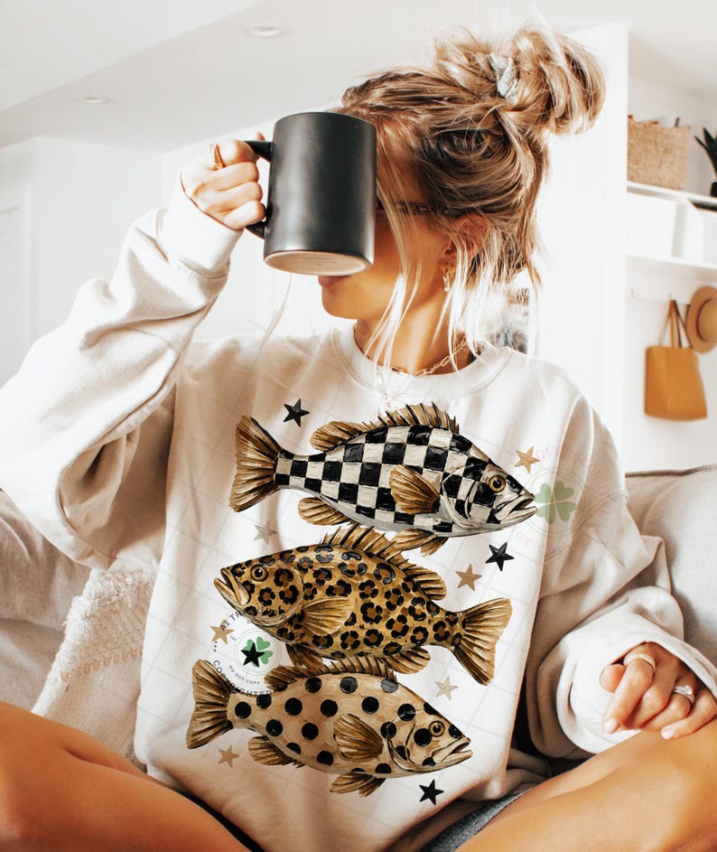 May include: Cream-colored sweatshirt featuring three fish designs: a black and white checkered fish, a leopard print fish, and a fish with brown polka dots. The sweatshirt also has star and clover graphics.