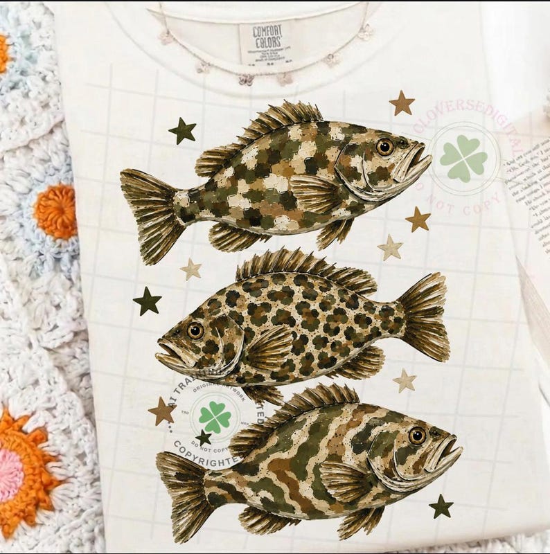 May include: White t-shirt with three detailed fish illustrations in shades of brown and green, each with a unique camouflage pattern. The shirt also features scattered brown stars and the text "COMFORT COLORS".