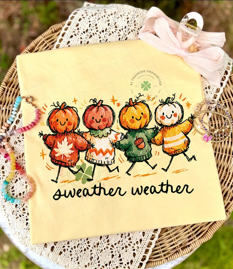 Może przedstawiać: Bladoż&oacute;łty t-shirt z grafiką przedstawiającą cztery dynie z kresk&oacute;wek o uśmiechniętych twarzach, z kt&oacute;rych każda ma na sobie sweter w innym kolorze. Dynie trzymają się za ręce. Pod ilustracją znajduje się napis "sweather weather".
