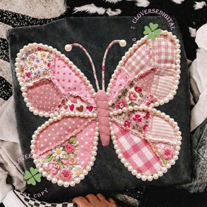 Może przedstawiać: Ozdobny motyl wykonany z różowych łatek materiału z kwiatowymi, groszkowymi i kratkowanymi wzorami. Motyl jest obrysowany sztucznymi perłami i ma haftowane detale. Tło to ciemnoszara tkanina. Widoczny jest tekst "CLOVERSE DIGITAL".