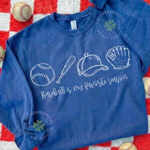 K&ouml;nnte beinhalten: Ein marineblaues Sweatshirt mit Baseball-Motiv. Das wei&szlig;e Design zeigt einen Baseball, einen Schl&auml;ger, eine Kappe und einen Handschuh mit dem Text "Baseball is my favorite season". Der Hintergrund ist ein rot-wei&szlig; kariertes Muster.