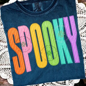 以下が含まれることがあります： ダークブルーの半袖Tシャツ。大きくカラフルな縦縞模様の文字で「SPOOKY」とプリントされています。各文字にはジグザグのステッチ風の縁取りが施されています。文字の色はオレンジ、ピンク、イエロー、グリーン、ティール、パープルです。