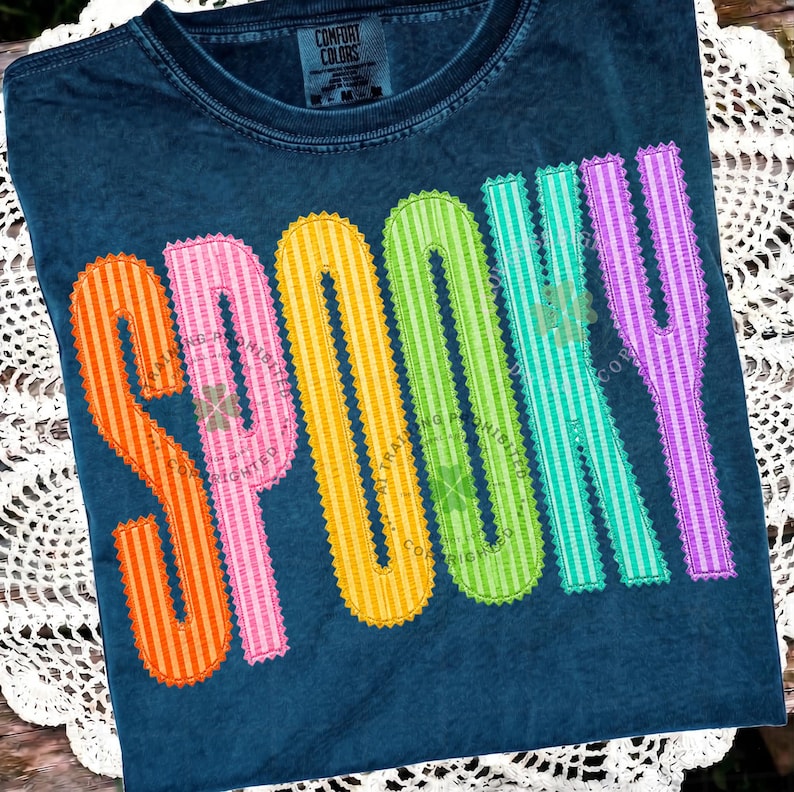 以下が含まれることがあります： ダークブルーの半袖Tシャツ。大きくカラフルな縦縞模様の文字で「SPOOKY」とプリントされています。各文字にはジグザグのステッチ風の縁取りが施されています。文字の色はオレンジ、ピンク、イエロー、グリーン、ティール、パープルです。