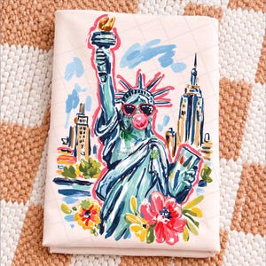 May include: A folded, light pink fabric featuring a colorful illustration of the Statue of Liberty wearing sunglasses and blowing a bubblegum bubble. The artwork includes New York City buildings and flowers.