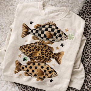 May include: Cream-colored sweatshirt featuring three whimsical fish designs. The top fish has a black and white checkered pattern, the middle fish has a leopard print, and the bottom fish has a polka dot pattern. Gold stars and a four-leaf clover accent the design.