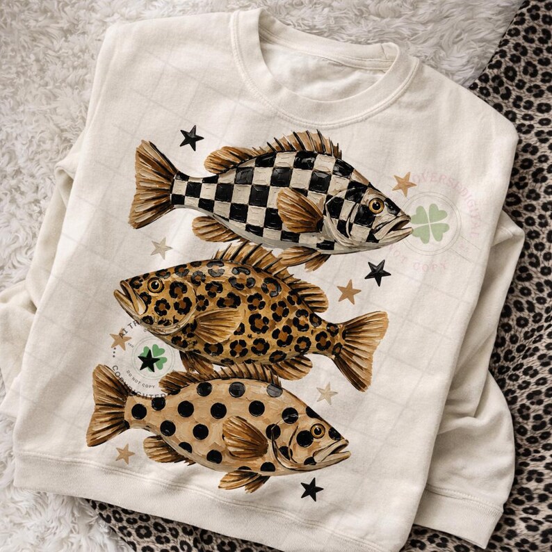 May include: Cream-colored sweatshirt featuring three whimsical fish designs. The top fish has a black and white checkered pattern, the middle fish has a leopard print, and the bottom fish has a polka dot pattern. Gold stars and a four-leaf clover accent the design.