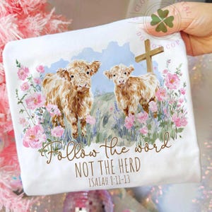 May include: White t-shirt featuring a watercolor illustration of two Highland cows amidst pink flowers and a wooden cross. The text "Follow the word NOT THE HERD" and Bible verse Isaiah 8:11-13 are printed below the image.