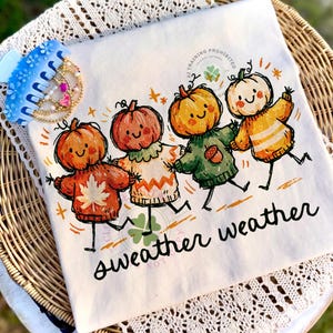 Może przedstawiać: Biały t-shirt z grafiką przedstawiającą cztery uśmiechnięte dynie, z kt&oacute;rych każda ma na sobie sweter w innym kolorze. Poniżej znajduje się napis "sweater weather" wykonany czarną kursywą oraz małe motywy liści i żołędzi.