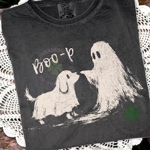 May include: A charcoal gray t-shirt featuring a distressed white graphic of a ghost and a dog wearing a ghost sheet. The text "Boo-p" is printed in a white script font above the dog. The shirt has a crew neckline and a Comfort Colors brand label.