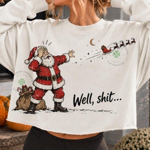 May include: A white long-sleeve cropped sweatshirt featuring a graphic of Santa Claus with a hand on his head, looking at a flying sleigh and reindeer in the distance. The text "Well, shit..." is printed in black script below the illustration.