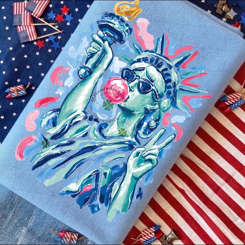 May include: A light blue towel featuring a whimsical illustration of the Statue of Liberty wearing sunglasses and blowing a bubblegum bubble. The artwork is in shades of blue, green, pink, and gold. The background includes American flags and patriotic decorations.