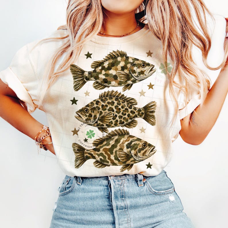 May include: Cream-colored t-shirt featuring three detailed fish illustrations with camouflage and leopard print patterns. The design includes star and shamrock accents, creating a unique and artistic aesthetic. A casual and stylish top.