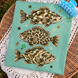 May include: A light teal fabric featuring three detailed fish illustrations with camouflage and leopard print patterns. Gold stars and text accents the design. A blue hair clip with floral details rests on the top right corner.