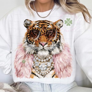 May include: White sweatshirt featuring a tiger wearing leopard print sunglasses, a pink feather boa, and a diamond necklace. The tiger's fur is orange and black. The sweatshirt has a clover logo.