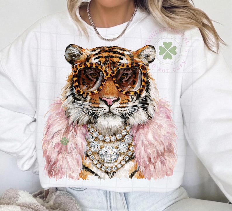 May include: White sweatshirt featuring a tiger wearing leopard print sunglasses, a pink feather boa, and a diamond necklace. The tiger's fur is orange and black. The sweatshirt has a clover logo.