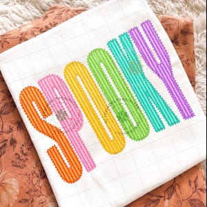 以下が含まれることがあります： 垂直の虹色のグラデーションでSPOOKYという文字が刺繍された白いTシャツ。各文字はオレンジ、ピンク、黄色、緑、ティール、紫などの異なる色で表現されており、ストライプ模様とスカラップ状の縁取りが施されています。