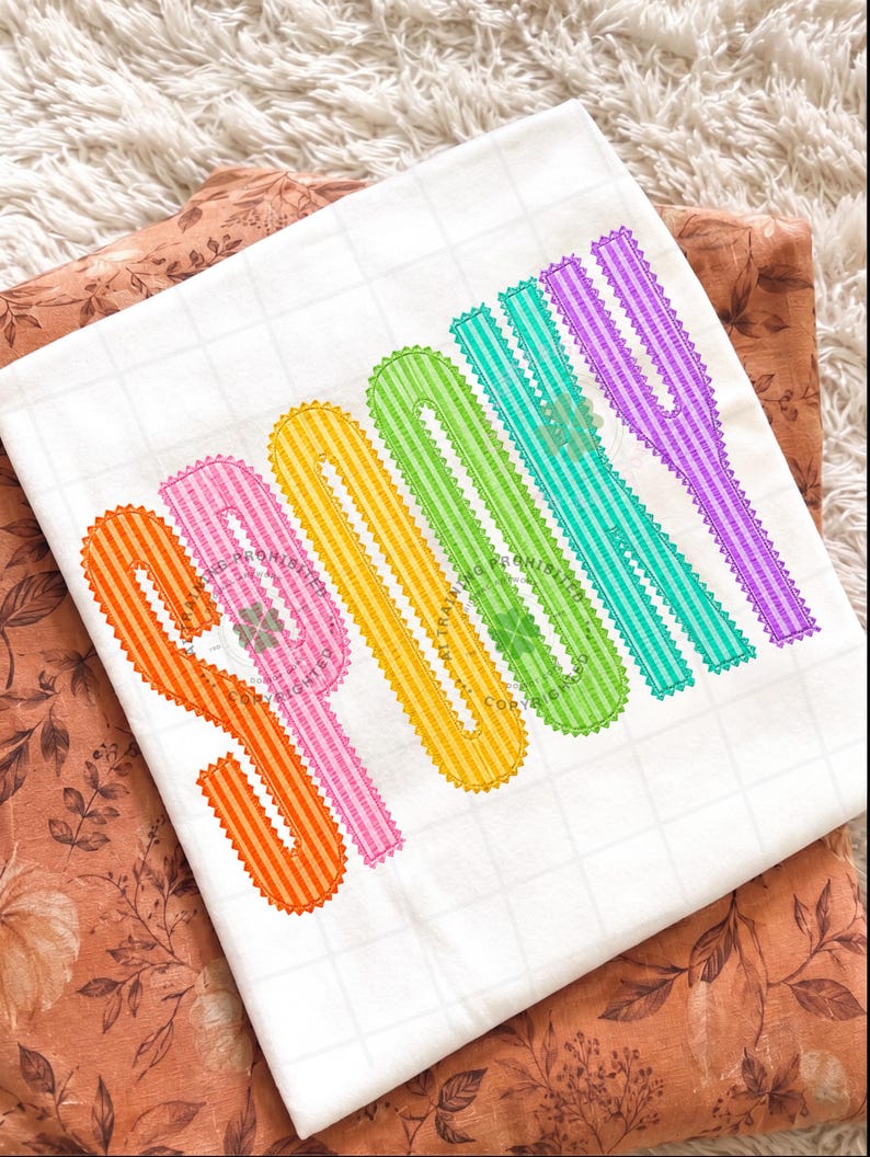 以下が含まれることがあります： 垂直の虹色のグラデーションでSPOOKYという文字が刺繍された白いTシャツ。各文字はオレンジ、ピンク、黄色、緑、ティール、紫などの異なる色で表現されており、ストライプ模様とスカラップ状の縁取りが施されています。