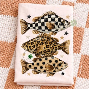 May include: A light pink decorative towel featuring three stylized fish. The top fish has a black and white checkered pattern, the middle fish has a leopard print, and the bottom fish has black polka dots. Gold and black stars are scattered around the fish.