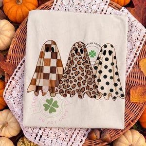 May include: A cream-colored t-shirt featuring a graphic of three ghosts. The first ghost has a brown and white checkered pattern, the second has a brown leopard print, and the third has black polka dots. Each ghost has two black oval eyes.