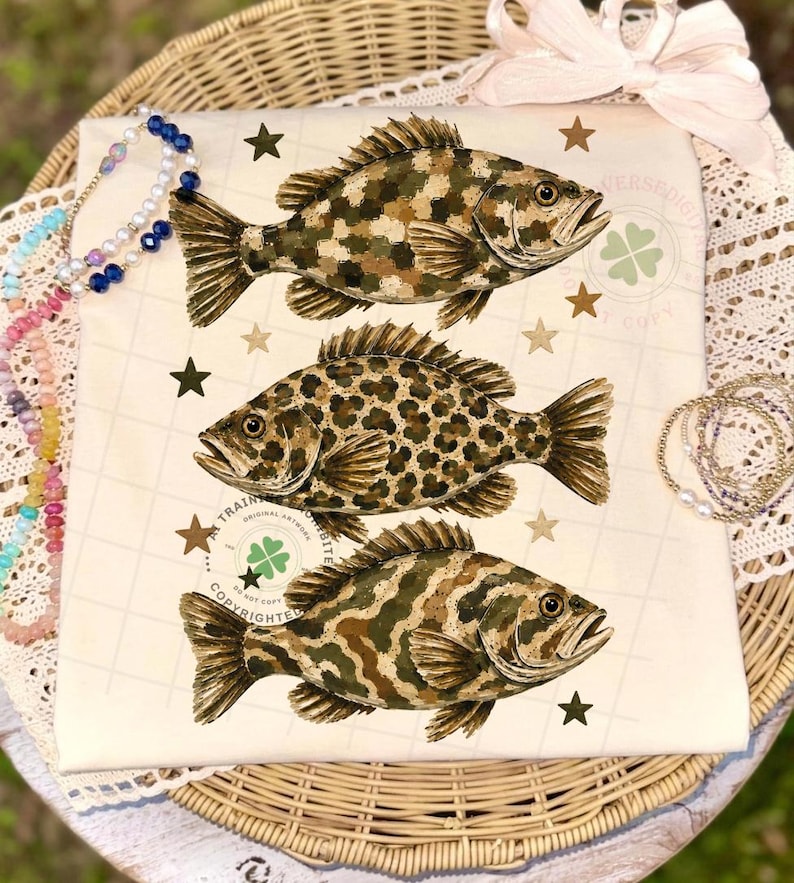 May include: Cream-colored fabric featuring three detailed fish illustrations with camouflage and leopard print patterns. The design includes brown stars and a clover emblem. The fabric is displayed on a wicker basket with decorative accessories.