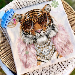 May include: A white square decorative pillow featuring a tiger wearing leopard print sunglasses, a pink feather boa, and a diamond necklace. The tiger has orange and black stripes. A blue hair clip with pearls is in the upper left corner.