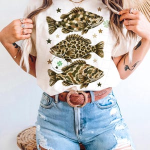 May include: Cream-colored t-shirt featuring three detailed fish illustrations in shades of brown and green, with star and clover accents. The shirt is paired with blue denim shorts and a brown belt with a heart-shaped buckle.