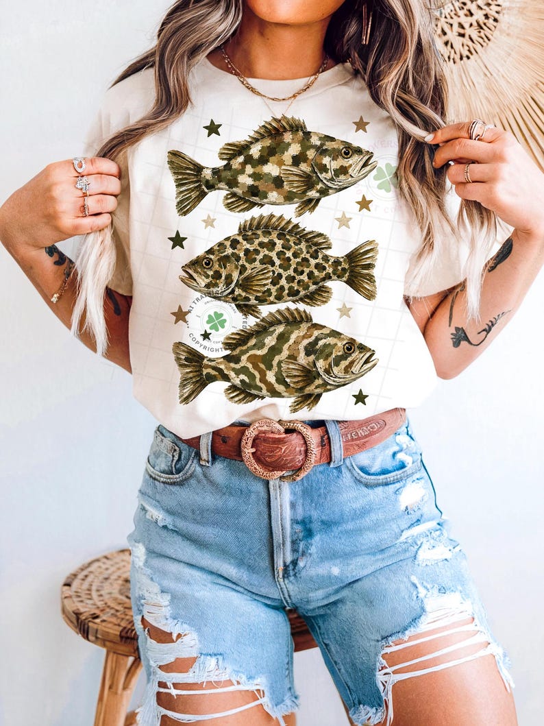 May include: Cream-colored t-shirt featuring three detailed fish illustrations in shades of brown and green, with star and clover accents. The shirt is paired with blue denim shorts and a brown belt with a heart-shaped buckle.