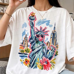 May include: White t-shirt featuring a colorful illustration of the Statue of Liberty wearing sunglasses and blowing a bubblegum bubble. The design includes New York City buildings and floral accents, creating a playful and artistic graphic.