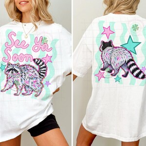 May include: White t-shirt featuring a colorful raccoon illustration with the text "See Ya Soon." The design includes pink stars and teal wavy lines. The back of the shirt has a similar design with a raccoon and stars.