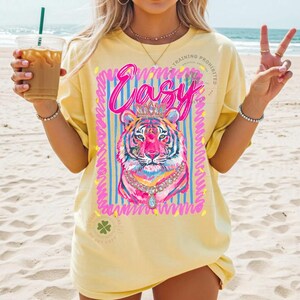 May include: Yellow t-shirt with a colorful tiger graphic wearing a crown and necklace. The word "Easy" is written above the tiger in pink script. The shirt has a relaxed fit and short sleeves.