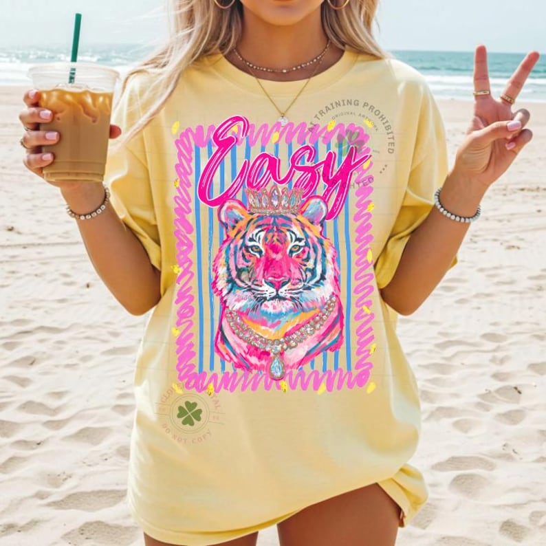 May include: Yellow t-shirt with a colorful tiger graphic wearing a crown and necklace. The word "Easy" is written above the tiger in pink script. The shirt has a relaxed fit and short sleeves.