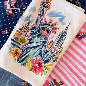 May include: White decorative towel featuring a colorful illustration of the Statue of Liberty wearing sunglasses and blowing a bubble. The artwork includes buildings, flowers, and a red outline. The towel is surrounded by patriotic decorations.