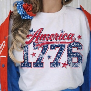 Może przedstawiać: Biała koszulka z napisem "America" w czerwonym skrypcie i numerem "1776" w kolorze niebieskim, z efektem postarzenia. Koszulka jest noszona z czerwoną, białą i niebieską kurtką.