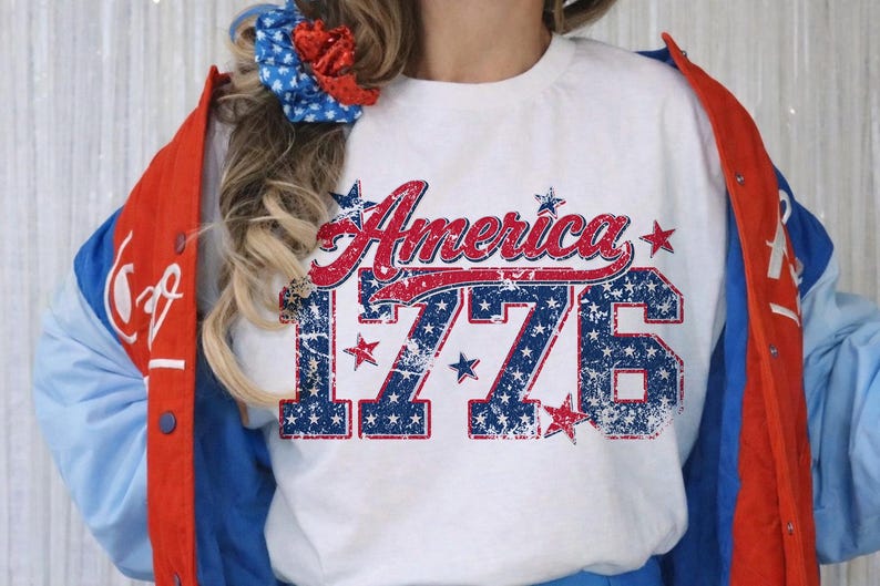 Może przedstawiać: Biała koszulka z napisem "America" w czerwonym skrypcie i numerem "1776" w kolorze niebieskim, z efektem postarzenia. Koszulka jest noszona z czerwoną, białą i niebieską kurtką.