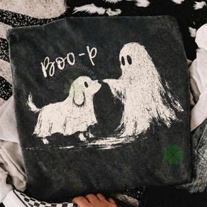 May include: A dark charcoal gray t-shirt featuring a white distressed graphic of a ghost and a dog wearing a ghost costume. The text "BOO-p" is printed in white above the figures. A small green four-leaf clover icon is visible in the bottom corner.