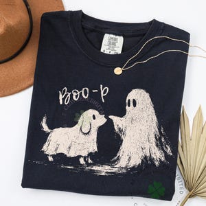May include: A dark navy blue crewneck t-shirt featuring a graphic print of a small dog and a ghost standing together. Above the figures, the text reads "BOO-p" in a handwritten style. The shirt has a Comfort Colors brand label at the neck.