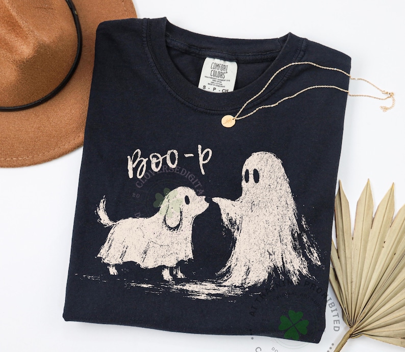May include: A dark navy blue crewneck t-shirt featuring a graphic print of a small dog and a ghost standing together. Above the figures, the text reads "BOO-p" in a handwritten style. The shirt has a Comfort Colors brand label at the neck.