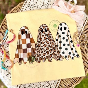 May include: A pale yellow t-shirt featuring a graphic of three ghosts. The first ghost has a brown and white checkerboard pattern, the second has a brown and black leopard print, and the third has black polka dots on a white background.