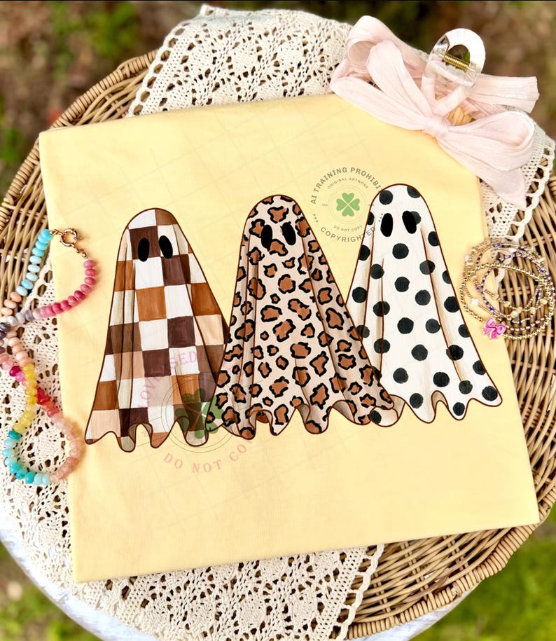May include: A pale yellow t-shirt featuring a graphic of three ghosts. The first ghost has a brown and white checkerboard pattern, the second has a brown and black leopard print, and the third has black polka dots on a white background.