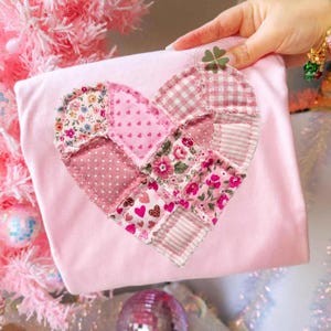 Könnte beinhalten: Ein hellrosa, quadratischer Gegenstand mit einem Patchwork-Herz-Design. Das Herz besteht aus verschiedenen rosa Stoffen, darunter Blumen-, Tupfen- und Streifenmuster. Der Gegenstand wird von einer Hand gehalten, im Hintergrund ist ein rosa Weihnachtsbaum zu sehen.