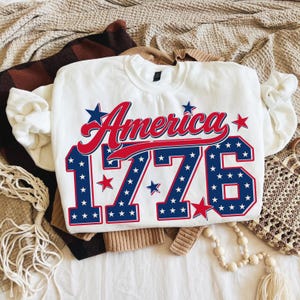 Może przedstawiać: Biała bluza z napisem "America" w czerwonym skrypcie i liczbami "1776" w kolorze niebieskim z białymi gwiazdami. Bluza jest złożona na wierzchu innych ubrań.