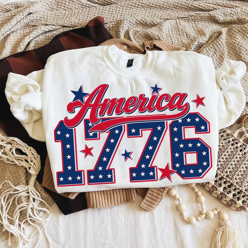 Może przedstawiać: Biała bluza z napisem "America" w czerwonym skrypcie i liczbami "1776" w kolorze niebieskim z białymi gwiazdami. Bluza jest złożona na wierzchu innych ubrań.