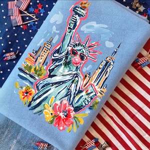 May include: A light blue decorative pillow featuring a whimsical illustration of the Statue of Liberty wearing sunglasses and blowing a bubble. The design includes colorful flowers and iconic New York City buildings. The pillow is surrounded by patriotic decorations.