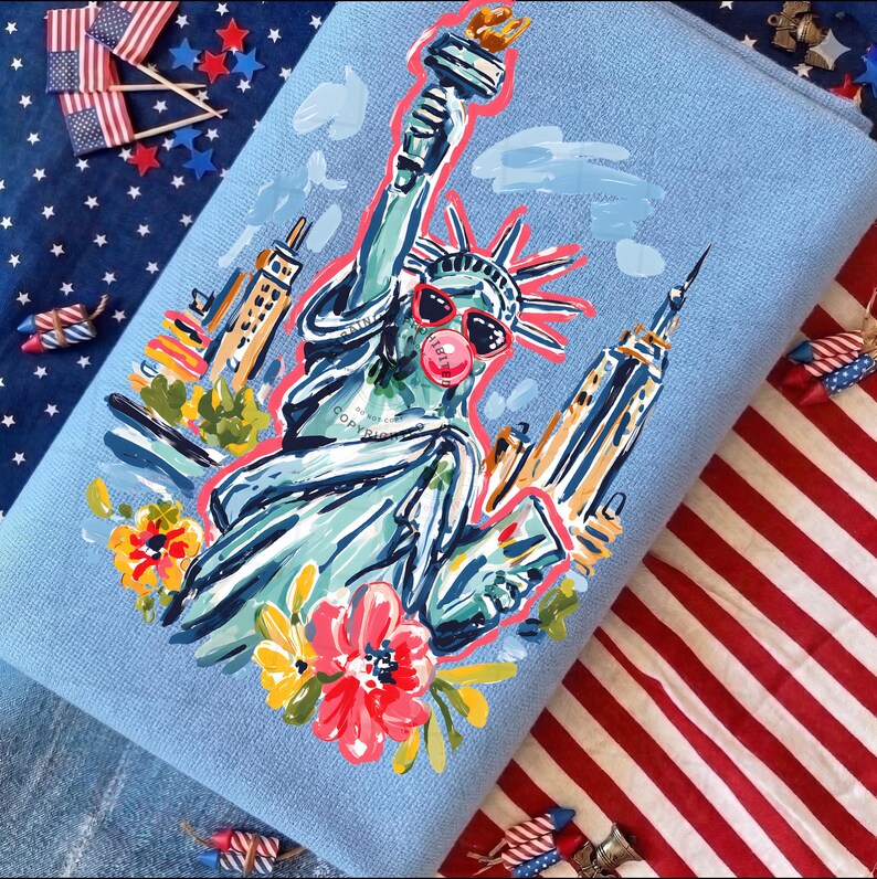 May include: A light blue decorative pillow featuring a whimsical illustration of the Statue of Liberty wearing sunglasses and blowing a bubble. The design includes colorful flowers and iconic New York City buildings. The pillow is surrounded by patriotic decorations.
