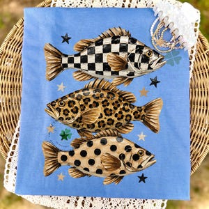 May include: A light blue decorative cloth featuring three stylized fish with unique patterns: a black and white checkerboard, a leopard print, and a spotted design. Black and gold stars are scattered around the fish. A white lace trim and a hair accessory are in the background.